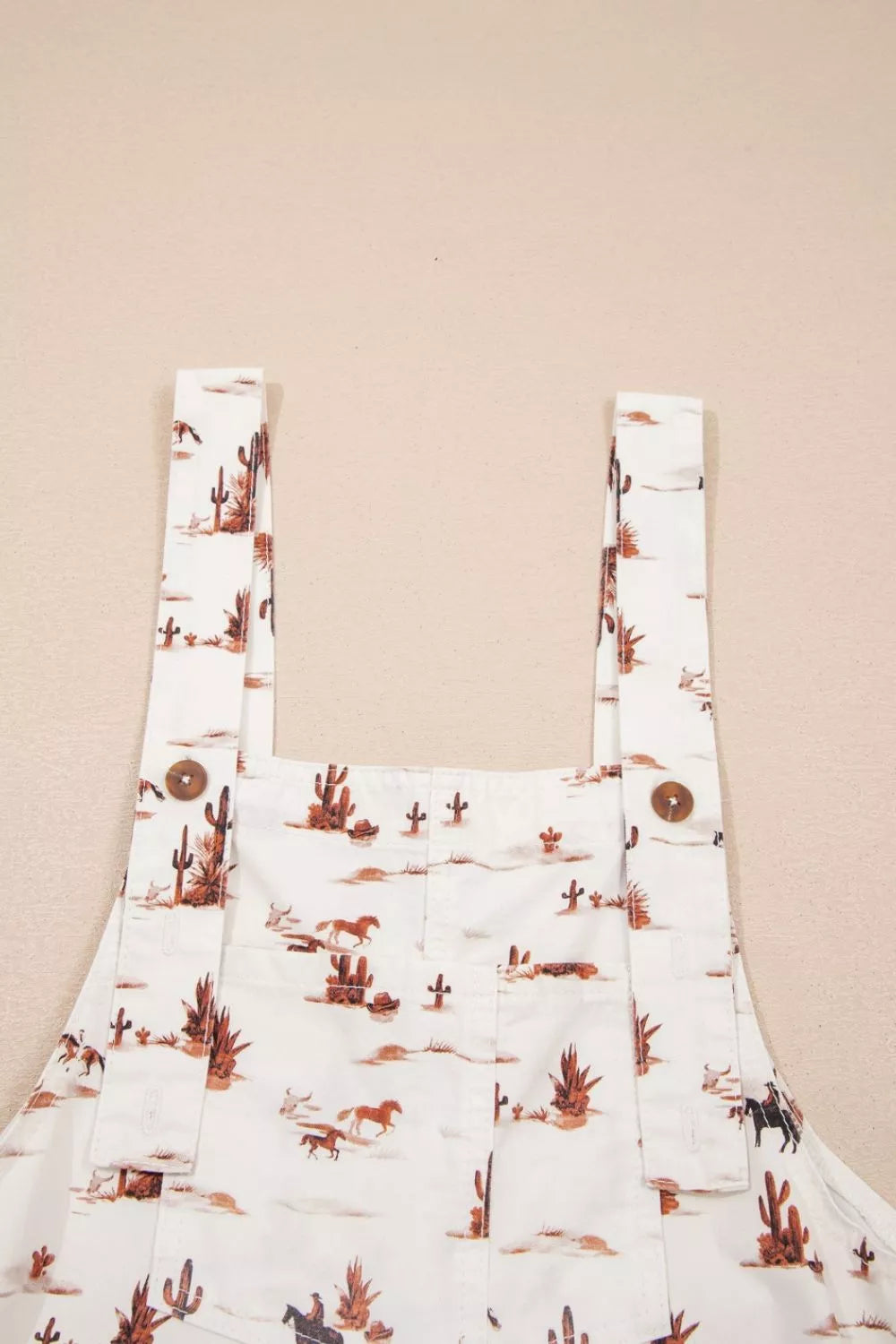 Front view of The Cowboy Way Overalls in cream with a Western desert print, adjustable straps, bib pocket, and wide legs.